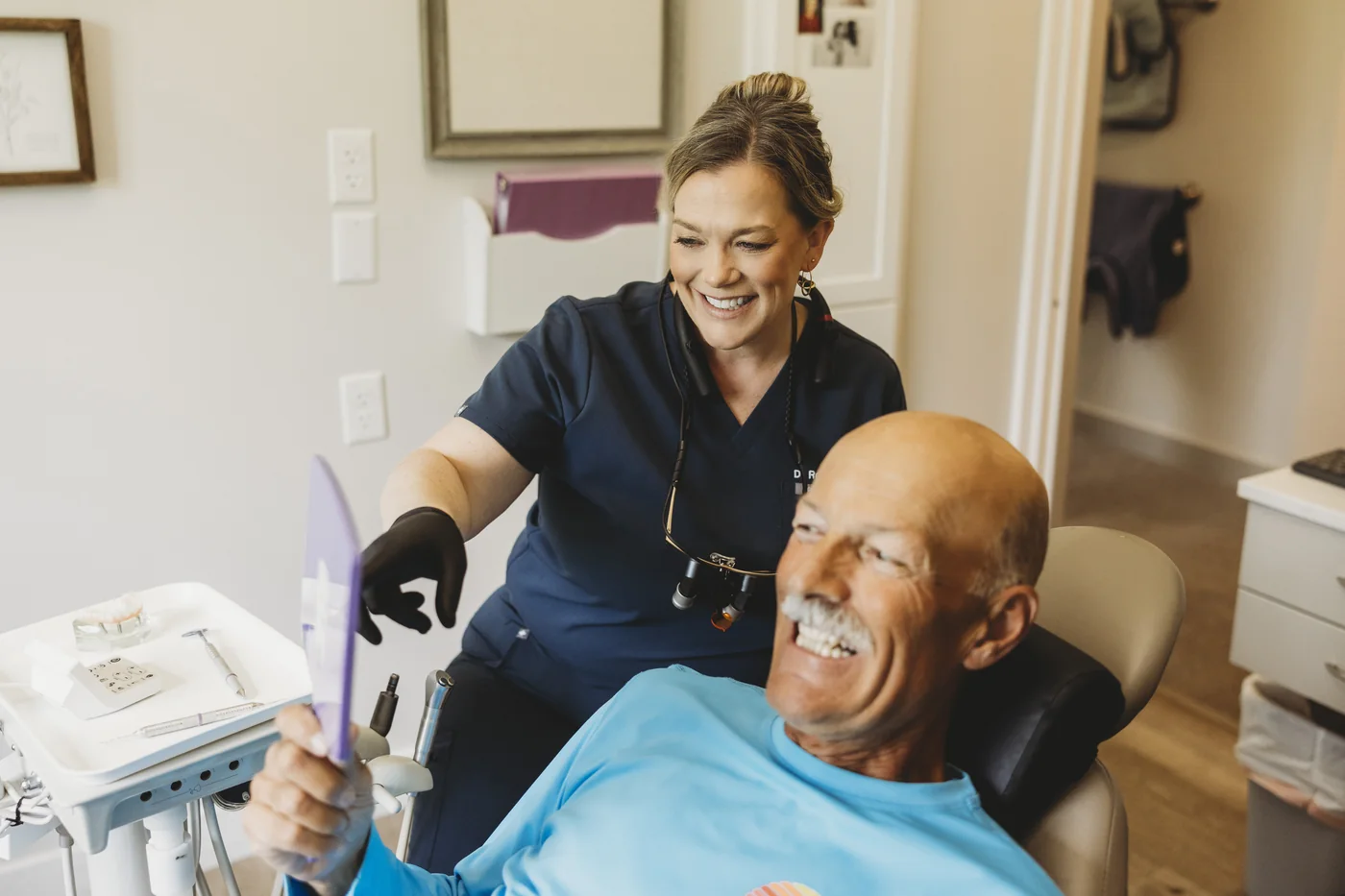 A patient smiling at his reflection in a hand mirror while his dentist looks on